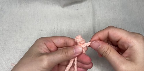 Hands forming a pink crochet frog knot closure, fingers holding the cord loops in position before tightening the knot. 