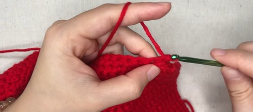 Hands using a green crochet hook to pick up stitches along the curved neckline edge of a red crochet fabric panel, showing the trim attachment step. 