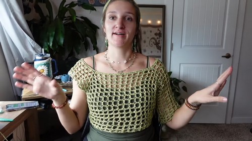 Tutorial creator wearing a sage green crochet mesh top with short sleeves, speaking to camera in a home studio with plants and artwork in the background 