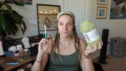 Tutorial creator holding a sage green yarn skein and a crochet hook, wearing a matching green headband in a home craft room 