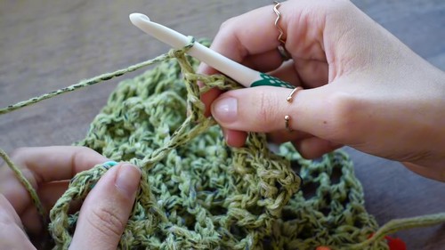 Hands crocheting green mesh fabric with a white hook, showing the open grid stitch in progress 