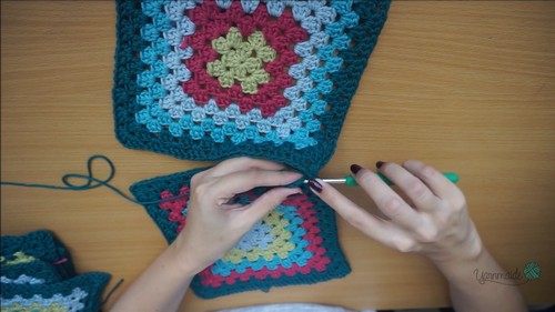 Two finished granny squares on a wooden surface, hands adding a new round with a crochet hook 