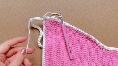 Hands working white picot edging along the top edge of a pink crochet bikini bottom with a pink hook