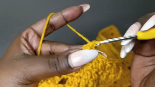 Close-up of hands working a crochet hook into a chain space in mustard yellow yarn, demonstrating a stitch step in the crochet shorts tutorial