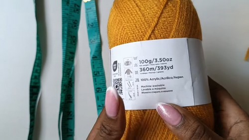 Skein of mustard yellow 100% acrylic yarn, 100g and 360 metres, held next to a tape measure 
