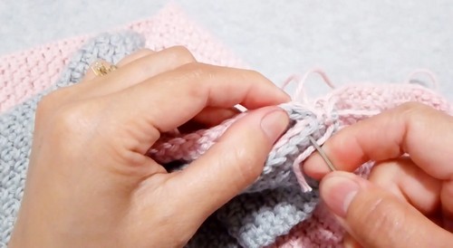 Close-up of hands seaming together two crochet panels in pink and gray-blue yarn with a tapestry needle, showing the joining step of the twist top construction 