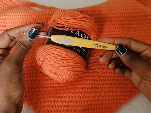 Hands holding a ball of orange yarn and a yellow 3.0 mm crochet hook resting on the finished fabric 