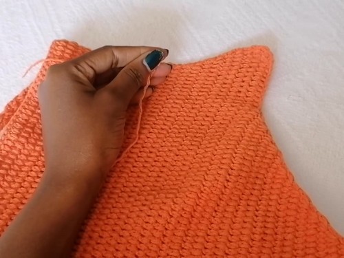 Hand holding the top edge of the orange crochet piece, weaving in a yarn end 