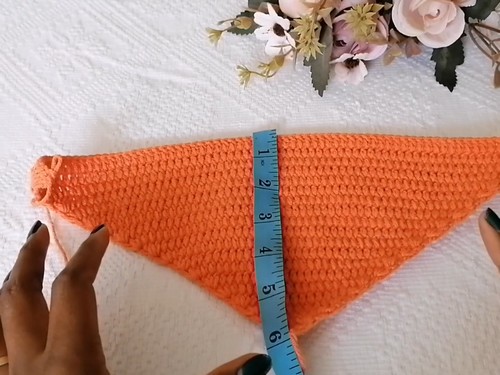 Orange triangular crochet piece laid flat with a measuring tape showing its depth, flowers in the background 