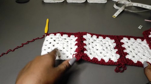 A long strip of joined red and white granny squares laid flat, with a hand pointing to the seam and a yellow hook resting nearby