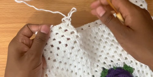Hands working the shoulder edge of the white granny stitch vest with a wooden hook