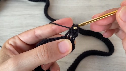 Hands crocheting into a black stitch with a gold hook, showing the dense single crochet texture up close
