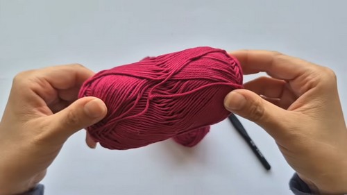 Hands holding a skein of dark red cotton yarn with a black crochet hook resting nearby 