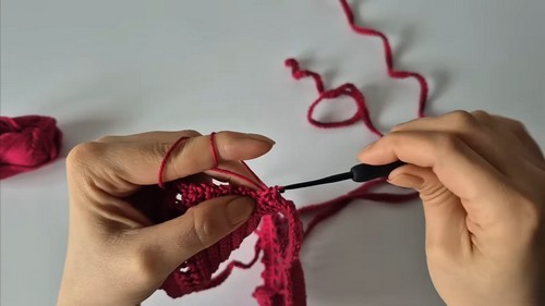 Close-up of hands crocheting a picot or shell edging along the curved edge of a red crochet bralette cup 