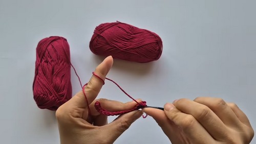 Hands forming a foundation chain with dark red yarn and a black crochet hook, two yarn skeins in background 