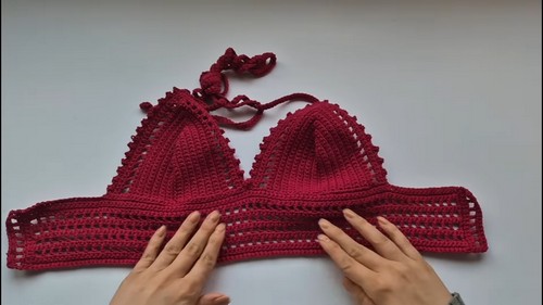 Hands smoothing a finished red crochet bralette laid flat, showing shaped cups and open underbust band detail 