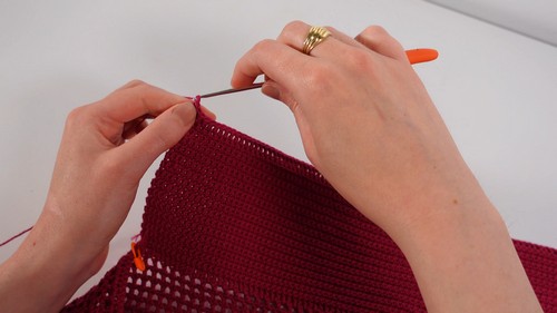 Hands working an orange crochet hook through the red fabric, joining or finishing a section of the solid and filet crochet top. 