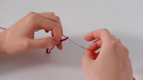 Hands forming an early chain or slip knot with red yarn and an orange crochet hook against a white background. 