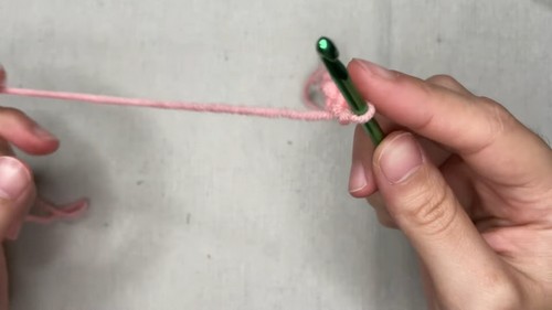 Hands holding a green crochet hook with pink yarn at a slip knot foundation 