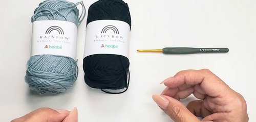Two skeins of Hobbii Rainbow cotton yarn in light blue and black beside a gold-tipped crochet hook on a white surface 
