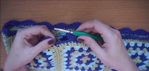 Hands crocheting a scalloped edge along the top of the granny square panel with a green hook
