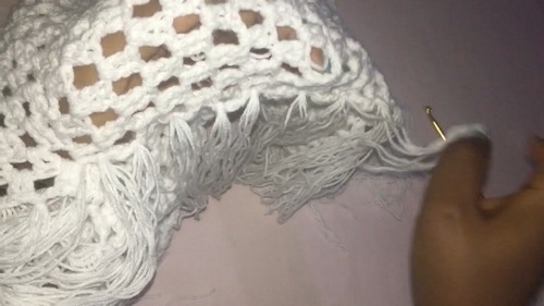 Hand attaching fringe strands to the hem edge of a white crochet mesh top with a gold hook 