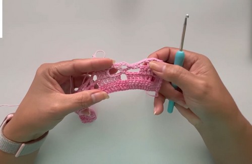 Hands working a pink crochet lace stitch with a blue hook, showing the open diamond repeat in progress 