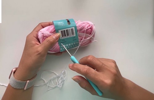 Hands holding a pink variegated yarn skein with label alongside a blue crochet hook 
