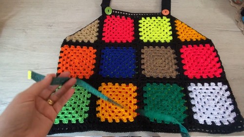 Completed crochet granny square patchwork top laid flat on a table, showing twelve multicolor squares in neon and earth tones with black border seaming and button straps 