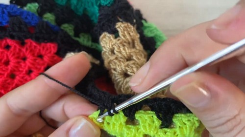 Hands joining two crochet granny squares with black yarn using a silver hook, working along the shared edge 