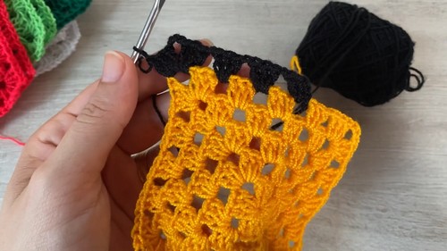 Hand holding a partially completed yellow granny square with black border in progress, silver hook and black yarn ball visible on a white surface 
