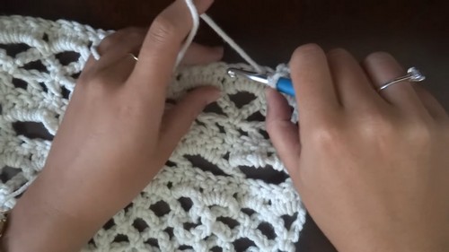 Hands working a blue crochet hook through a cream lace panel, showing the open shell stitch pattern in the fabric up close. 