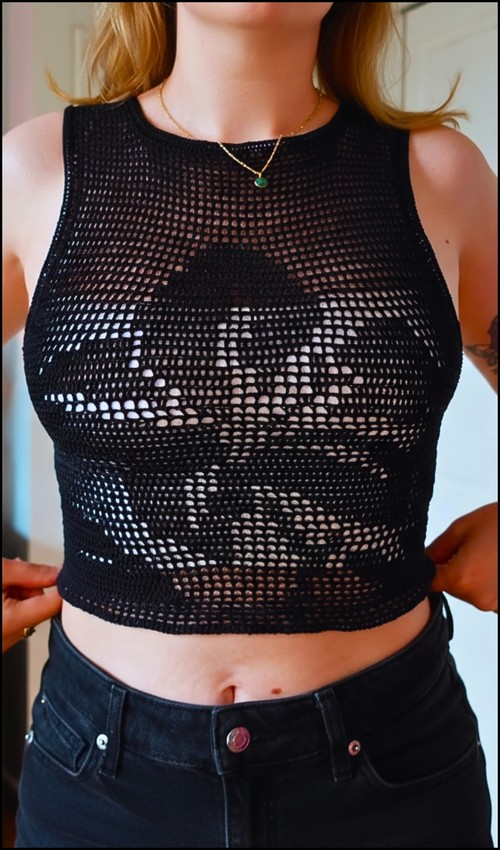 Black filet crochet crop tank top on a model, featuring a large flower motif formed by filled and open double crochet squares across the front, worn over black jeans with a gold pendant necklace. 