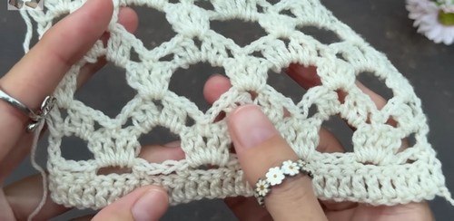Hands holding a cream crochet swatch showing a repeating shell lace stitch pattern with open arches, fingers spreading the fabric to show the stitch structure.
