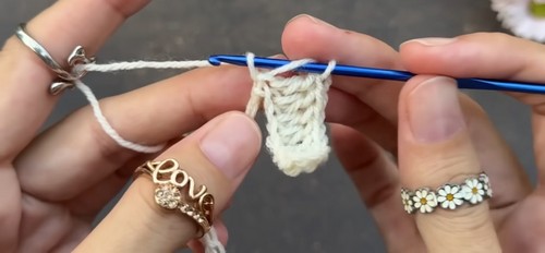 Hands forming the beginning stitches of a cream crochet project with a blue hook, showing the first shell cluster taking shape on the foundation chain.