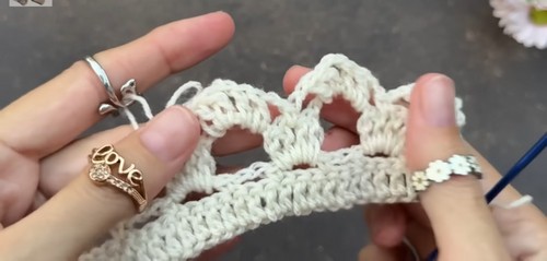 Hands working a blue crochet hook through a cream shell lace swatch, showing the placement of stitches within the open arch of the previous row.