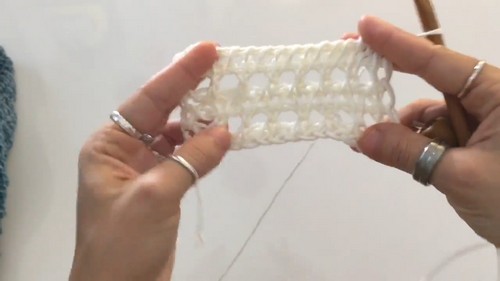 Hands holding a white Tunisian crochet swatch with a long Tunisian hook, demonstrating the open elongated-loop mesh stitch structure used throughout the blouse.
