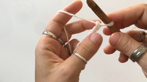 Hands tensioning white yarn across the fingers with a Tunisian crochet hook, showing the setup position for working Tunisian simple stitch rows.