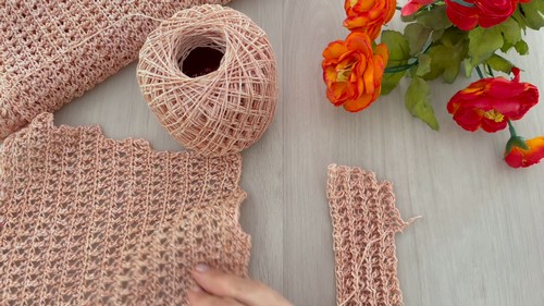 Flat lay of shaped blush crochet trouser leg panels beside a ball of linen yarn and orange flowers