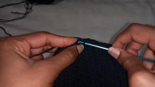 Image 3 — Hands working a thin metal hook through the edge of a dark navy crochet piece, showing stitch placement along a finished row. 