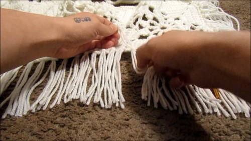 Hands pulling and separating long cream fringe strands along the hem of a crochet boho top to finish the fringe detail