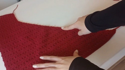 Hands smoothing a large flat red crochet body panel on a white surface, showing even stitch rows and armhole shaping at the top edge 