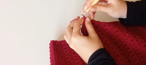 Close-up of hands working a bamboo crochet hook into red crochet fabric at the shoulder or armhole area