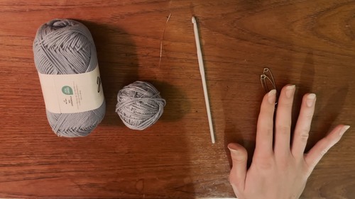 Two balls of grey yarn and a wooden crochet hook laid out on a wooden surface alongside a hand for scale, showing the materials used for the project. 