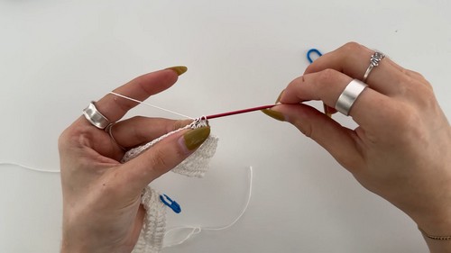 Hands working early chain stitches in white yarn with a red crochet hook, with stitch markers visible on the work surface. 