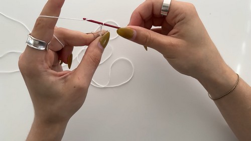 Hands forming the very first slip knot or foundation loop in white yarn with a red crochet hook. 