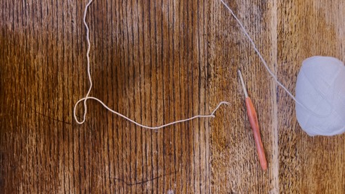 A wooden crochet hook, a ball of light grey yarn, and a length of starting chain laid out on a wooden table, showing the materials used for the mesh top.