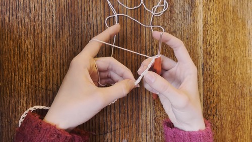 Hands forming a slip knot with fine white yarn on a wooden table, beginning the foundation chain for a mesh crochet project. 