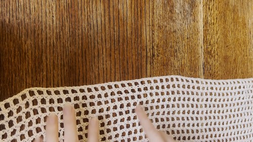 Hands spreading a section of cream crochet mesh fabric on a wooden surface, showing the even square grid of the mesh stitch pattern. 