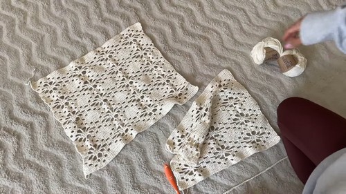 Three cream crochet panels — one large body piece and two smaller sleeve sections — laid out on a textured surface beside a yarn ball, showing the garment pieces before joining